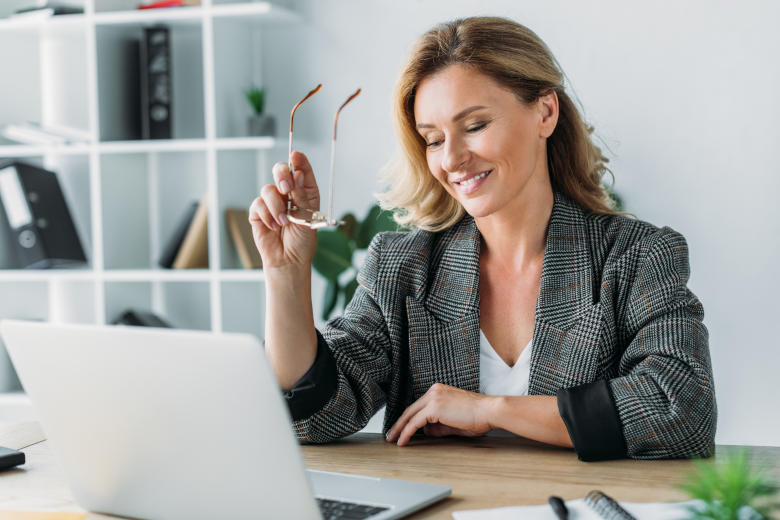 Frau_schaut_Website_am_Laptop Eine lächelnde Frau sitzt bei Ihrem Schreibtisch mit abgesetzter Brille und schaut auf Ihrem Laptop Ihre Website an.